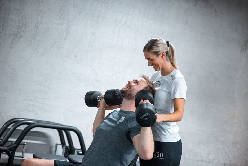FITGO's personlige trener som hjelper mann med å trene med dumbbells mens han sitter på benk og tar skulderpress